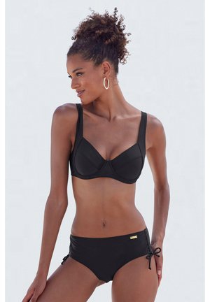 Göndör hajú nő, fekete bikini felsővel, merevítővel és megkötős alsóval, balra néz, fehér háttér előtt.