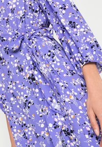 Kukallinen mekko kevyestä purppurakankaasta, jossa on mustat, valkoiset, vaaleanpunaiset ja oranssit kukkamaalaukset, vyötäröllä solmittava yksityiskohta ja puhvelihihat.