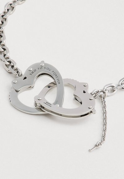 Strieborný kovový náramok srdcového tvaru, ktorý má zámky v tvare srdca a je na ňom vyryté "NAVŽDY PRIPOJENÝ K TEBE", spojený hrubým retiazkou.