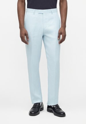 Mężczyzna w jasno niebieskich, dopasowanych spodniach, czarnych skórzanych mokasynach i ciemnogranatowej koszuli na jednolitym białym tle.