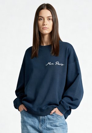 Donna in piedi con le mani nelle tasche dei jeans, indossa una felpa blu navy morbida con la scritta "Mia Paris" sul petto su uno sfondo neutro.