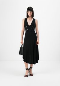 Mustapuku, jossa on syvä V-skirt, istuva bodice, jossa on kiiltävä, rakenteellinen korostus. Laskeutuva hame ja nilkkaketjulliset korkokengät. Pieni musta käsilaukku.