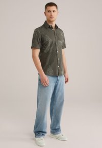 Camisa de manga corta de color verde oscuro con patrones de hojas blancas, cuello abotonado, combinada con jeans rectos de color azul claro y zapatillas blancas.