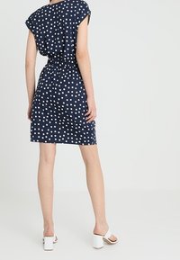 Navyblaues Kleid mit weißen Punkten, kurzen Ärmeln und geraffter Taille, das eine lockere Passform und einen knielangen Saum hat. Getragen mit weißen Absatzsandalen.