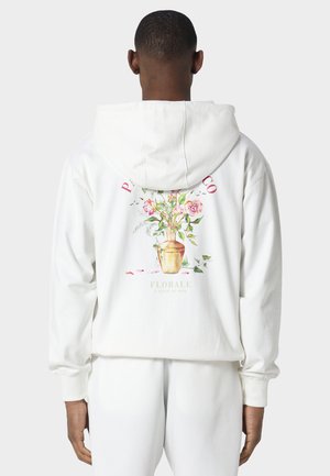 Hombre que lleva sudadera blanca con capucha y pantalones, con gráfico de jarrón floral colorido y texto "Florale" en la parte trasera de la sudadera.