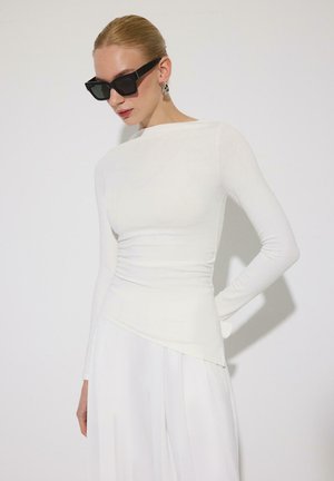 Femeie cu păr blond prins într-un coc elegant poartă ochelari de soare negri supradimensionați, cercei argintii, un top alb mulat cu mânecă lungă și pantaloni albi cu picioare largi.