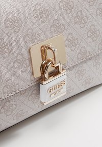 Beżowa, teksturowana torebka projektanta z prostokątnym zapięciem w kolorze złotym oraz dołączoną białą kłódką z grawerem "GUESS EST. 1981."