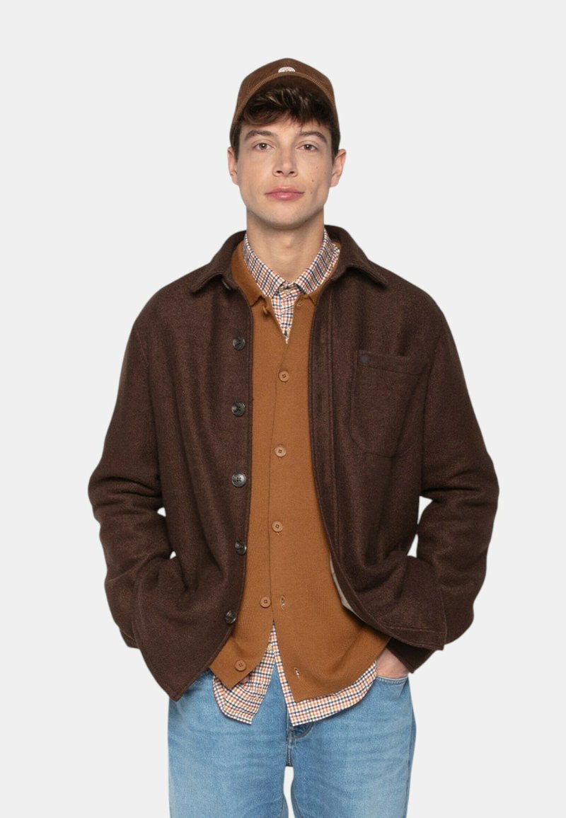 Homme portant une casquette de baseball marron, une veste boutonnée marron foncé sur un cardigan beige et une chemise à carreaux, les mains dans les poches d'un jean bleu clair.