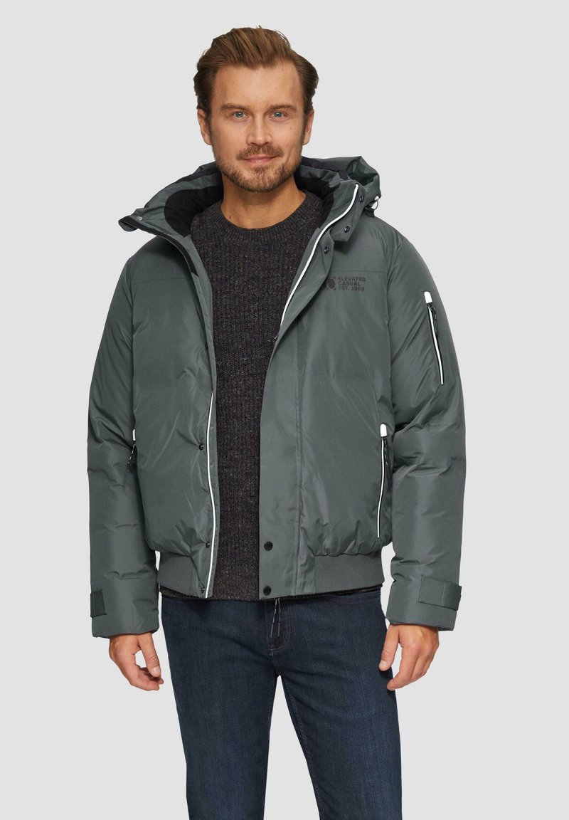 Ciemnozielona kurtka puchowa z kapturem, z białymi zamkami błyskawicznymi, ściągaczami na rękawach oraz logo na lewej piersi. Noszona na szarej bluzie.