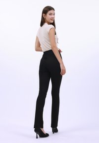 Pantaloni neri aderenti con vita alta, abbinati a una maglietta beige con maniche a cappuccio. Il modello indossa scarpe con tacco nere, in piedi su uno sfondo bianco.