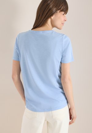 Femme aux cheveux bruns portant une chemise à manches courtes bleu clair et un pantalon blanc, légèrement tournée vers la droite sur un fond uni.