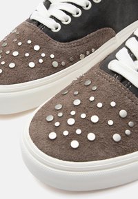 Brune semskede sneakers med hvite snøring, med metalliske nagler i forskjellige størrelser på tåboksen og en kontrasterende mørk overdel. Gummisåle.