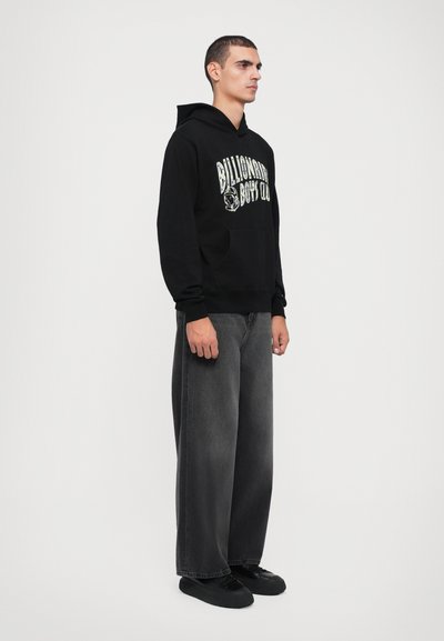 Sweatshirt à capuche noir avec le grand texte blanc "Billionaire Boys Club" sur le devant ; porté avec un jean ample et foncé et des chaussures noires.