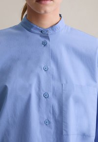 Lichtblauw overhemd met knoopsluiting en een mandarin-kragen, voorzien van zes bijpassende knopen en een borstzak aan de linkerzijde. Gladde stoftextuur.