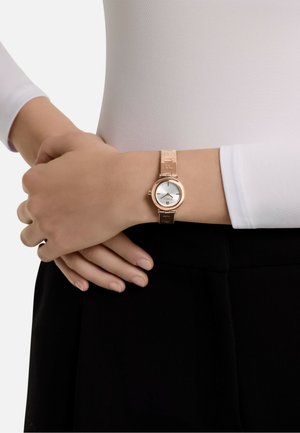 Orologio rosa oro con cassa rotonda, quadrante bianco e bracciale in metallo a motivi. Indossato a un polso con una maglietta attillata di colore chiaro.