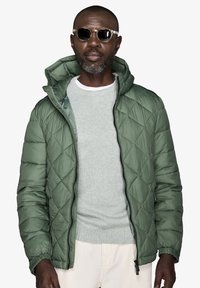 Vatteret, grøn pufferjakke med hætte, der har lynlås, elastiske manchetter og en let glans, båret over en grå sweater.