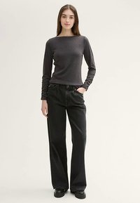 Grijs shirt met lange mouwen en een vierkante halslijn, gecombineerd met zwarte jeans met wijde pijpen. Model staat met handen in de zakken en draagt zwarte schoenen.