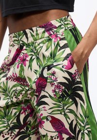 Persona con pantalones de estampado tropical con pájaros rosas y hojas verdes, mano descansando en el bolsillo, y top corto negro.