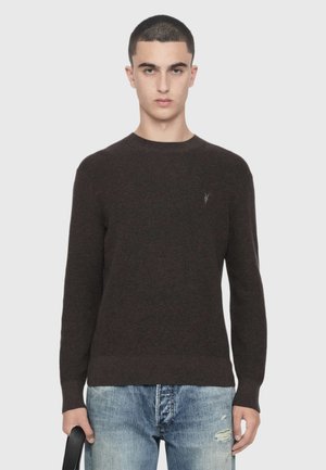 Ung mand med kort hår iført en mørkebrun sweater og lyseblå slidte jeans stående mod en ensfarvet lys baggrund.