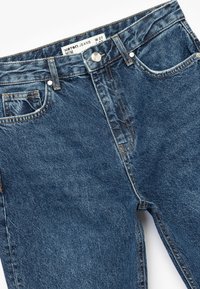Jeans de mezclilla azul oscuro con talle alto, que cuentan con un cierre de botón, detalles de pespuntes y dos bolsillos frontales. Etiqueta de tamaño: W 27.