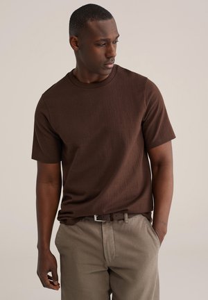 Bruin T-shirt met korte mouwen en een geribbelde textuur, voorzien van een ronde hals en een losse pasvorm, gecombineerd met beige broek en een bruine riem.