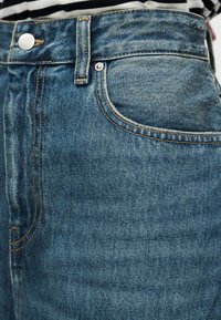 Τζιν παντελόνι με ψηλή μέση σε μπλε χρώμα, με ξεθωριασμένη υφή, αντίθετη ραφή, μπροστινή τσέπη και κλείσιμο με μεταλλικό κουμπί στη μέση.