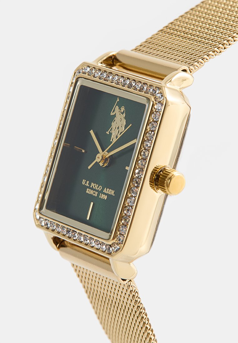 Montre rectangulaire en or avec un cadran vert, ornée de cristaux autour du visage, et un bracelet en mailles métalliques. Présente une couronne en or texturée.
