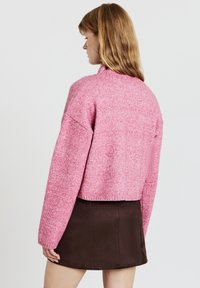 Rosa gestrickter Pullover mit hohem Kragen und überschnittenen Schultern, kombiniert mit einem braunen Rock. Die Textur wirkt weich, mit einem gesprenkelten Muster.