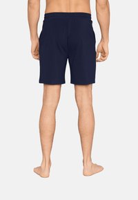 Mies, joka pukeutuu tummansiniset keskipitkät shortsit, näkyy takaa, seisoo paljain jaloin yksinkertaista taustaa vasten.