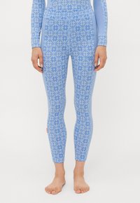 Pernas em leggings azuis justas com um padrão floral geométrico branco, de pé descalças contra um fundo simples.