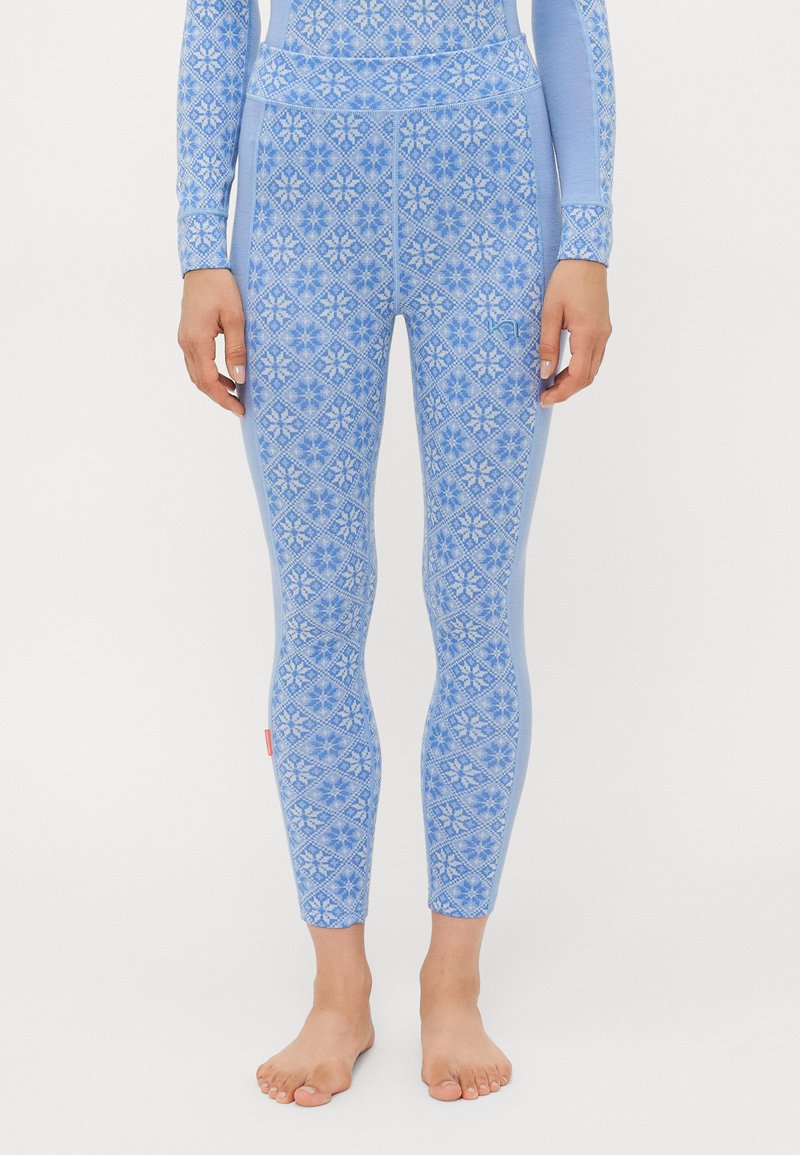 Pernas em leggings azuis justas com um padrão floral geométrico branco, de pé descalças contra um fundo simples.