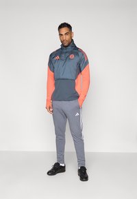 Sportsoutfit bestående af en grå og lys orange pullover med lynlås, lange ærmer, og grå bukser med tre hvide striber på siden.