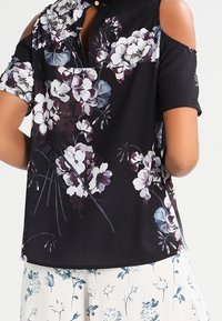 Blusa negra con estampado floral en blanco, morado y azul. Presenta un diseño de hombros al aire, mangas cortas y un cuello con detalle de botón.