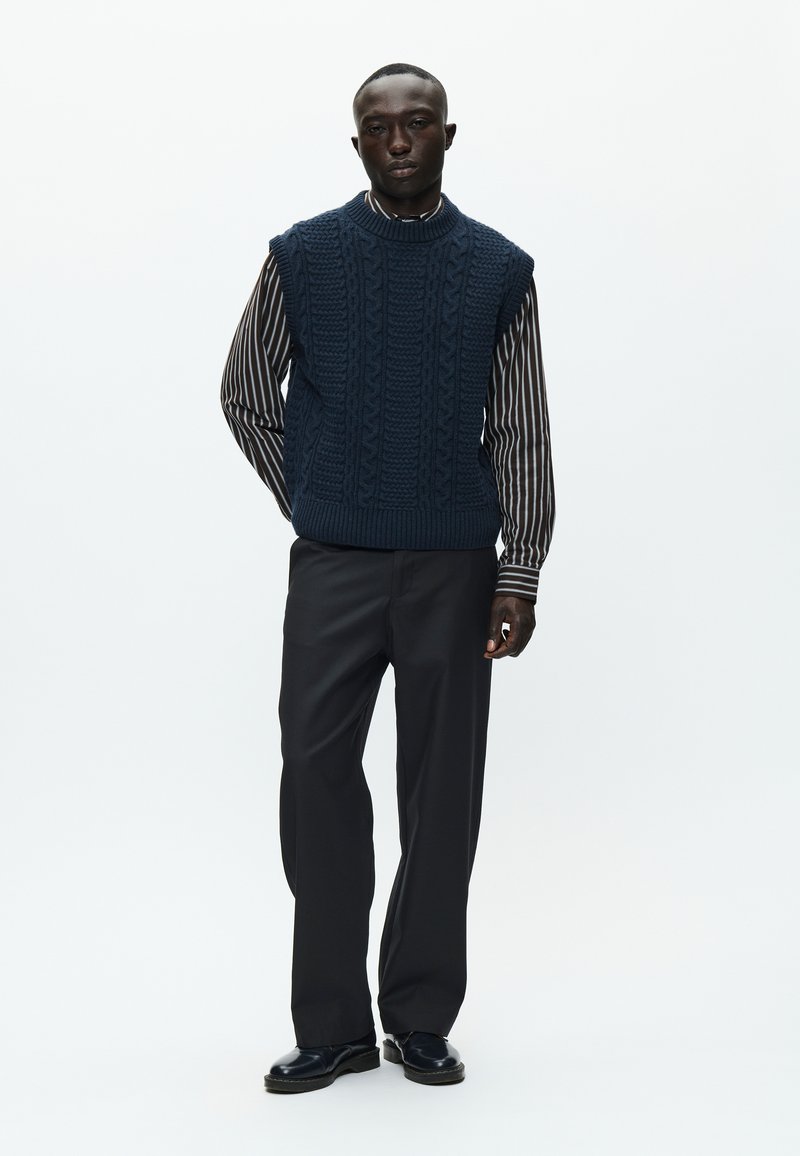 Pull en laine sans manches bleu marine avec un motif en relief, porté par-dessus une chemise à manches longues noire et blanche à rayures, associé à un pantalon sombre et des chaussures.