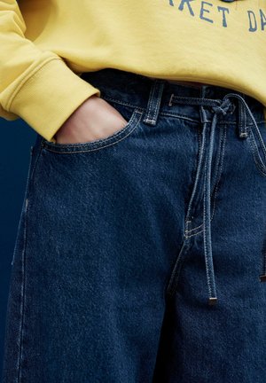 Dark blue denim jeans with a drawstring waist, featuring open side pockets and light stitching details. Worn with a yellow sweatshirt.