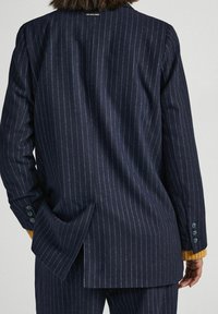 Woman wearing a navy pinstripe blazer with back vent and matching trousers, with a mustard-yellow cuffed sweater underneath.