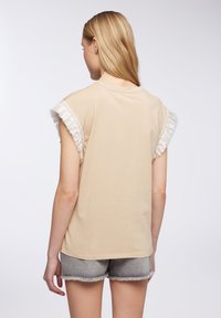 Donna con lungo capelli biondi, che indossa un top senza maniche beige con dettagli in frange bianche e pantaloni corti in denim grigio, di spalle rispetto alla fotocamera.