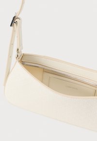 Crème-kleurige handtas met een textuur buitenkant en een geometrisch patroon, met een ritsluiting en een verstelbare schouderband. Het interieur heeft een kaartvak.