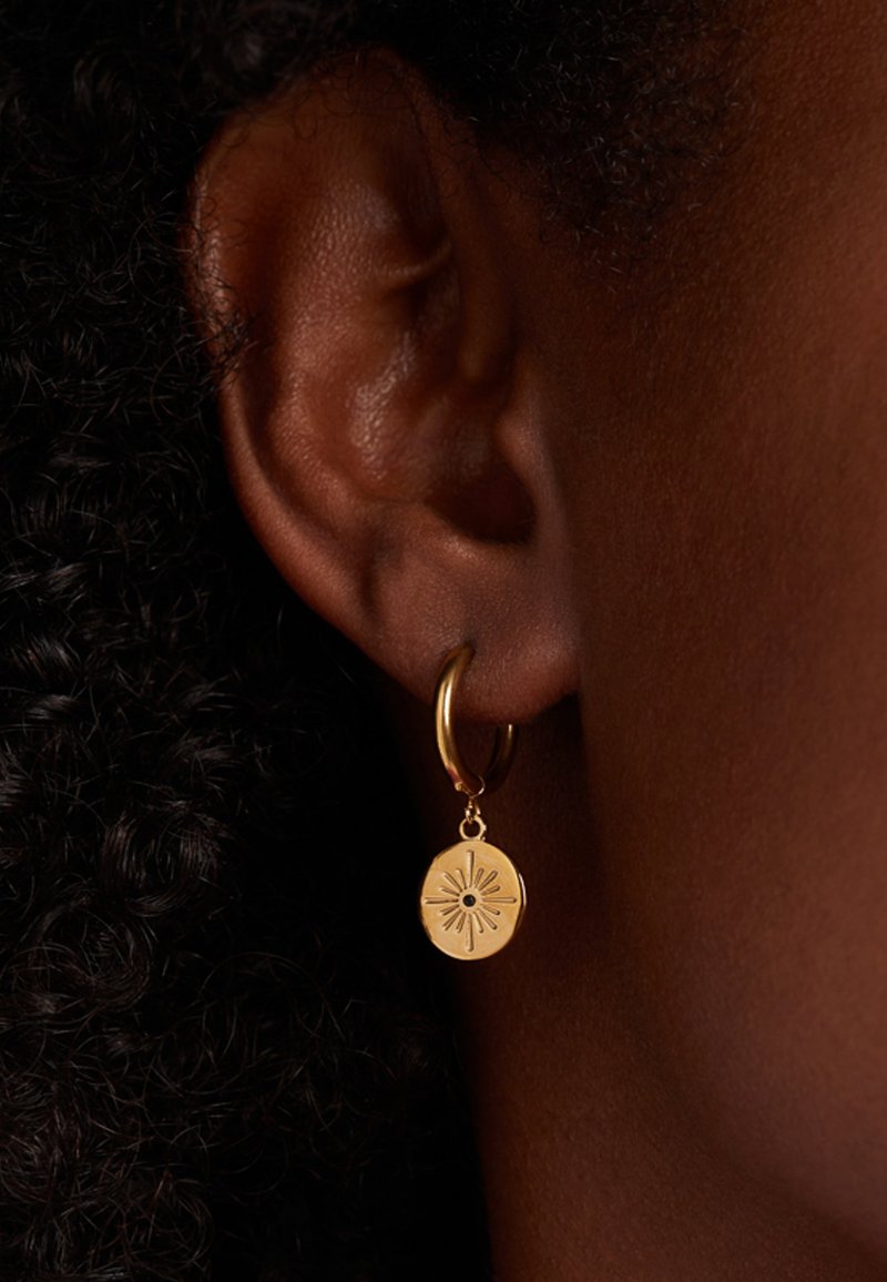 Boucle d'oreille en or avec un charme circulaire orné d'un motif de rayons de soleil et d'un point central noir. La vue en gros plan met en évidence la texture et les détails.