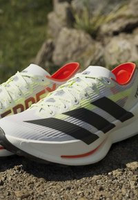 Weiße und schwarze Laufschuhe mit neongelben Schnürsenkeln und orangefarbenem Innenfutter, im Freien auf Kies mit verschwommenen Steinen im Hintergrund platziert.