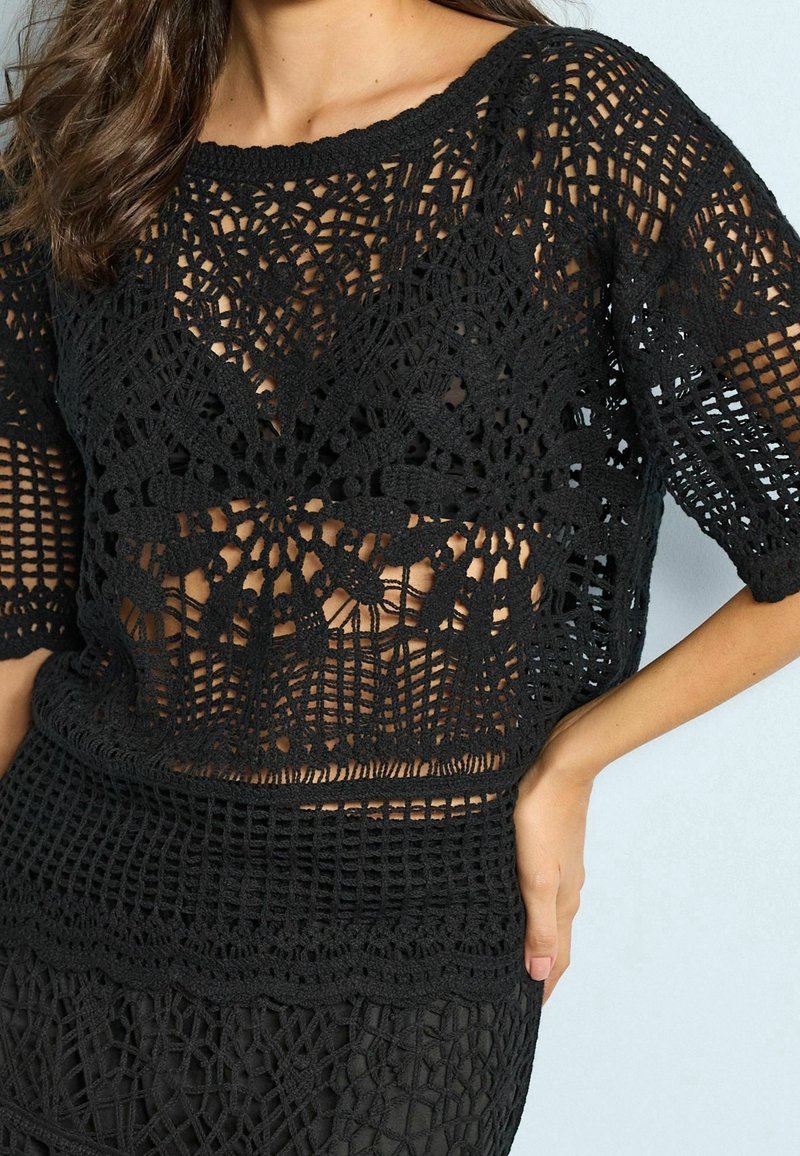 Haut noir en crochet avec des motifs géométriques et floraux, manches mi-longues et détails semi-transparents, porté par une personne avec une main sur la hanche.