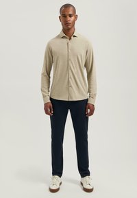 Beige overhemd met lange mouwen, kraag en knoopsluiting, gecombineerd met marineblauwe broek en witte schoenen. Gladde textuur, minimaal ontwerp.