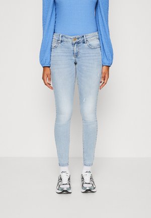 Osoba ubrana w jasnoniebieskie obcisłe dżinsy, niebieską teksturowaną bluzkę z długim rękawem, białe skarpetki i szaro-białe trampki, stojąca na jednolitym tle.