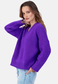Pull en maille violet à col en V avec poignets et ourlet côtelés, présentant une coupe ample et une texture douce, associé à un jean bleu clair déchiré.