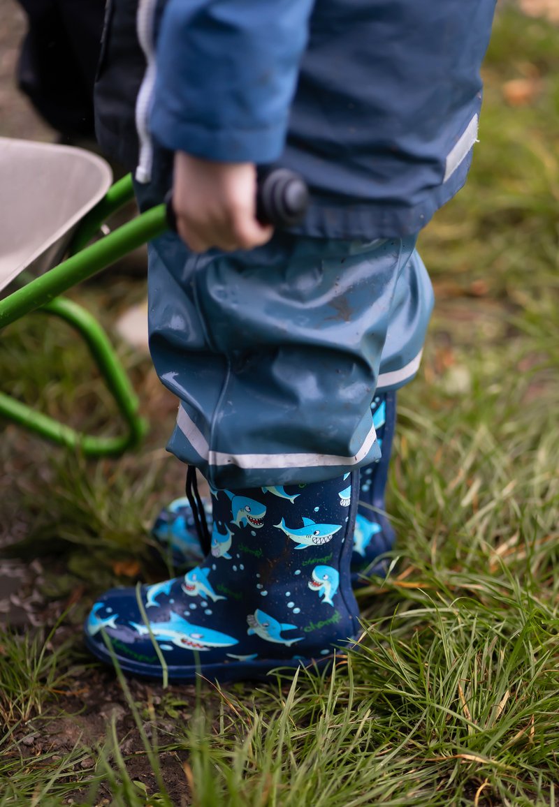 Bottes de pluie bleues avec des motifs de requin, portées avec un pantalon imperméable bleu clair. Une brouette verte à proximité sur l'herbe.