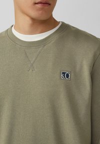 Uomo che indossa un felpa verde oliva con un piccolo logo ricamato "s.O" sul petto, sovrapposta a una maglietta a maniche corte bianca a coste.