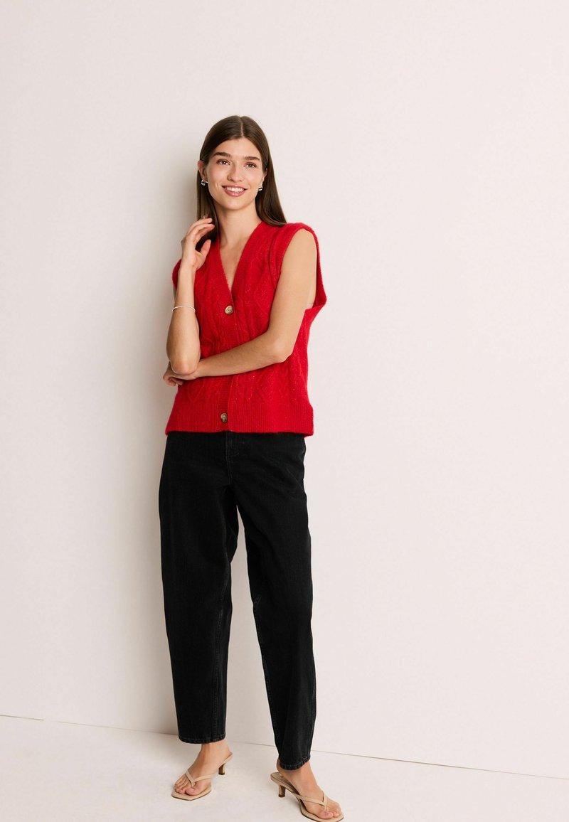 Gilet rosso lavorato a maglia con scollatura a V profonda, maniche corte e dettaglio con bottoni, abbinato a jeans neri larghi e sandali con tacco color castano.