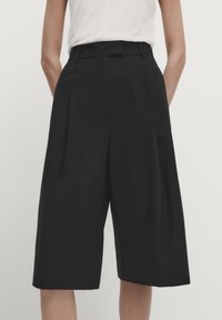 Musta culottes-housut, joissa on laskokset, korkeavyötärö, sileä kangas ja sivutaskut. Polvenpituinen ja rento istuvuus, yhdistetty valkoiseen yläosaan.