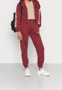 Burgundin vihreä vetoketjullinen huppari, jossa valkoista tekstiä, yhdistettynä saman sävyisiin verryttelyhousuihin. Asusteena valkoiset urheilukengät. Mukana reppu.