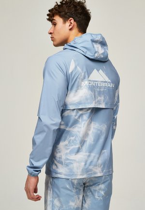 Mand iført blå og hvide tie-dye jakke og bukser med hætte, set fra siden, der viser "Monterrain Mountain Exploration" logo på ryggen.